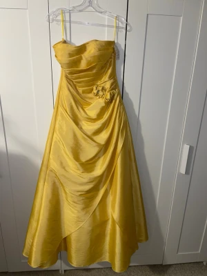 Gul balklänning  - Elegant gul långklänning med draperad framsida. Klänningen har dekorativa blommor med glittrande stenar i midjan och ett glansigt material som ger en lyxig känsla. Perfekt för speciella tillfällen. Snörning bak i ryggen för förbättrad passform.