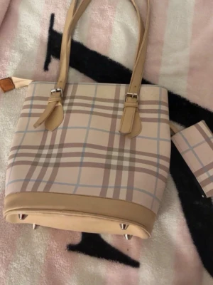 Beige-rutig handväska med plånbok - Snygg handväska i beige och ljusbrunt rutigt mönster med matchande plånbok. Väskan har två långa handtag, silverfärgade detaljer och förstärkt botten. Perfekt för dig som gillar klassisk och stilren design.