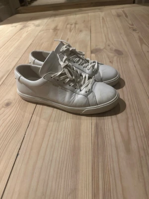 Vita Saint Laurent sneakers i läder - Klassiska vita sneakers från Saint Laurent i mjukt läder med diskret logga i guld på sidan. Modellen har lågt skaft, vita snören och en stilren, minimalistisk design som passar till det mesta. Perfekta för dig som gillar exklusiva och tidlösa skor.