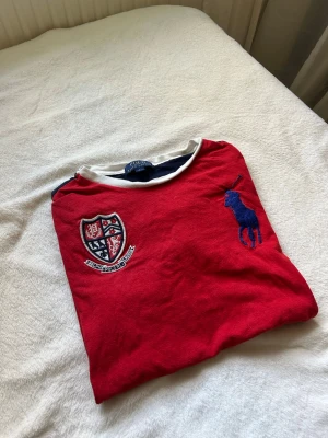 Röd t-shirt från Polo Ralph Lauren - Röd t-shirt från Polo Ralph Lauren med marinblå ärmar och vit krage. Stor blå logga på bröstet och ett broderat klubbmärke på vänster sida. Klassisk passform och mjukt bomullsmaterial, perfekt till jeans eller shorts.