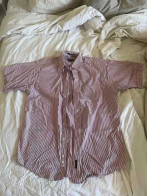 Randig kortärmad skjorta från Gant storlek M - Säljer en snygg kortärmad skjorta från Gant i vitt och rött med tunna vertikala ränder. Skjortan har klassisk krage, knappar framtill och en relaxed passform. Perfekt för dig som gillar stilrena och tidlösa plagg. Storlek M