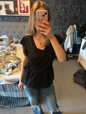 Svart omlottopp med volangärm - Snygg svart omlottopp med volangärmar och knytband i midjan. Toppen har en djupare ringning och ett mjukt, lätt tyg som faller fint. Perfekt att styla med jeans eller kjol för en trendig look. Mycket fin och aldrig använd!