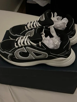 Dior B30  - Snygga svarta Dior sneakers med vita och silverfärgade detaljer. Skorna har en låg profil, vita snören och en tjock vit sula med Dior-logga på sidan. Materialet är en mix av textil och syntet, och designen har en modern streetwear vibe.