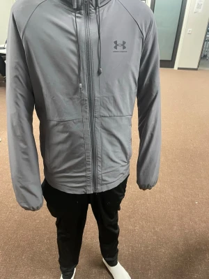 Grå vindjacka från Under Armour - Snygg grå vindjacka från Under Armour med dragkedja framtill och justerbar huva med dragsko. Jackan har elastiska muddar vid ärmsluten och en minimalistisk design med logga på bröstet. Perfekt för blåsiga dagar och enkel att matcha med olika outfits.