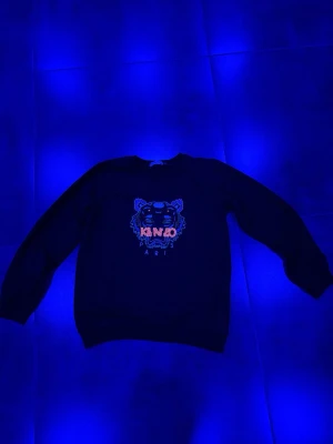 Svart Kenzo sweatshirt med tigertryck - Cool svart sweatshirt från Kenzo med det ikoniska tigerhuvudet och loggan i orange och blått framtill. Tröjan har rund hals, långa ärmar och ribbade muddar. Perfekt för dig som gillar streetwear och vill sticka ut med en snygg detalj.