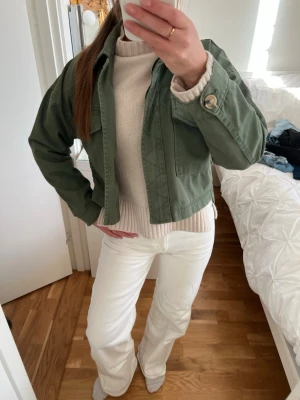 Grön croppad jeansjacka - Snygg croppad jeansjacka i militärgrönt med stora bröstfickor och knappdetaljer vid ärmslut. Jackan har en avslappnad passform och är tillverkad i bomull, perfekt att slänga över en tröja för en casual look.