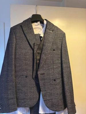 Grå rutig kostym med väst från HM - Snygg grå rutig kostym från H&M med matchande väst och kavaj. Kavajen har två knappar, fickor och ett paisleymönstrat innerfoder. Västen är mörkgrå med fyra knappar och två fickor. Perfekt för dig som vill ha en stilren och modern look.