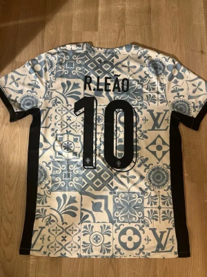 Portugal Louis Vuitton X Nike Tröja - Säljer en unik Portugal LV X Nike fotbollströja med R. Leão och nummer 10 på ryggen. Tröjan har ett blåvitt kakel-inspirerat mönster, svarta sidopaneler och korta ärmar. Perfekt för dig som vill sticka ut under sommaren🌞