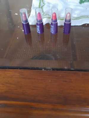 Fyra mini läppstift i olika färger - Säljer ett set med fyra mini läppstift i olika nyanser: röd, rosa, korall och nude. Alla har lila hylsor och genomskinliga lock. Perfekta att ha med i väskan för snabba touch-ups. Läppstiften har klassisk rundad form och ger en snygg finish.