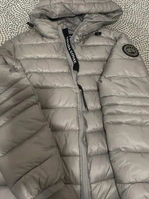 Ljusgrå pufferjacka från Canada Goose - Snygg ljusgrå pufferjacka från Canada Goose med huva och dragkedja framtill. Jackan har det klassiska Canada Goose-märket på ärmen och är tillverkad i ett glansigt, quiltat material som håller värmen under kalla vinterdagar.