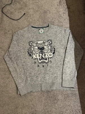 Grå sweatshirt från Kenzo med tiger - Grå sweatshirt från Kenzo med det ikoniska tigermotivet och texten 'KENZO PARIS' broderat på bröstet. Tröjan har rund hals, långa ärmar och ribbade muddar. Perfekt för dig som gillar streetwear och vill sticka ut med en klassisk design.