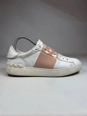 Rosa Valentino Garvani Open - Snygga vita sneakers från Valentino med en bred, glansig rosa rem över snörningen och nitar på hälen. Skorna har en klassisk låg siluett och är tillverkade i läder med gummisula. Perfekta för dig som gillar stilrena men ändå unika detaljer.