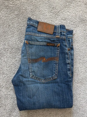 Blå jeans från Nudie Jeans - Säljer ett par klassiska blå jeans från Nudie Jeans med snygga slitningar och kontrastsömmar. Modellen har raka ben och har en brun läderpatch bak i midjan. Materialet är mjukt denim i bomull och passformen är slim. 