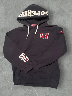 Unik NY Superdry hoodie - Svart hoodie från Superdry med stor huva och vita dragsnören. På bröstet finns ett rött NY-tryck och på huvan står det SUPDRY i stora bokstäver. Ärmen har även ett broderat SD-tryck. Hoodien är i mjuk bomullsblandning och har känguruficka framtill.