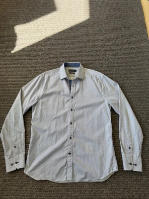 Skjorta - Snygg skjorta från Riley med tunna vertikala ränder i vitt och blått. Klassisk krage, långa ärmar och mörka knappar framtill. Tillverkad i bomull för en skön känsla och stilren look.