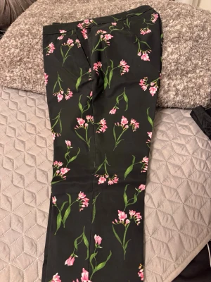 Svarta byxor med blommigt mönster - Snygga svarta byxor med rosa och grönt blommönster. Byxorna har rak passform och är perfekta för dig som vill sticka ut lite extra. Materialet känns mjukt och har en lätt glans. Midjan är normal och modellen är klassisk med dragkedja och knapp.
