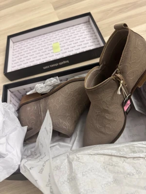 Beige stövletter från Bugatti - Snygga beige stövletter från Bugatti med låg klack och diskret mönstrad yta. Skorna har dragkedja på insidan och en liten metallogo på sidan. Tillverkade i syntetmaterial med träfärgad sula. Perfekta för dig som vill ha en stilren och trendig look.