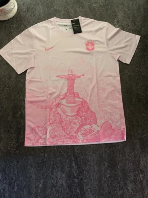 Rosa Nike t-shirt med Rio-motiv - Snygg ljusrosa t-shirt från Nike med korta ärmar och tryck av Kristusstatyn i Rio de Janeiro framtill. T-shirten har Nike-logga och ett emblem på bröstet i matchande rosa toner. Perfekt för dig som gillar unika prints och streetwear vibes.