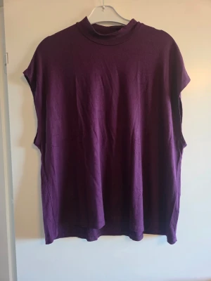 Lila ärmlös topp, stl XL  - En enkel och snygg lila topp med lös passform och ärmlös design. Toppen har en hög krage och är tillverkad i mjukt material, perfekt för att styla lager på lager eller bära som den är.