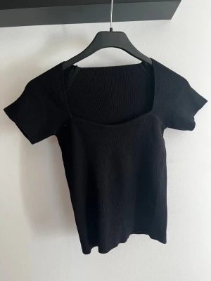 Svart ribbad topp med fyrkantig ringning - En svart ribbad topp med korta ärmar och fyrkantig ringning. Toppen har en figurnära passform och är tillverkad i ett stretchigt material som ger en bekväm känsla. Perfekt att styla med jeans eller kjol. Aldrig använd 