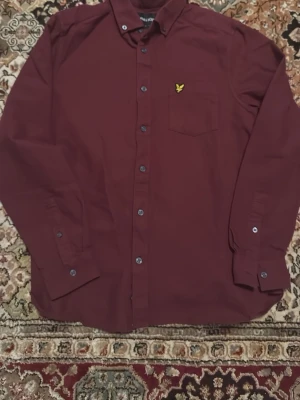 Vinröd skjorta från Lyle & Scott - Snygg vinröd skjorta från Lyle & Scott med klassisk krage och knappstängning framtill. Bröstficka med gul broderad logga. Skjortan har långa ärmar och en clean, stilren look som passar till många outfits. Storlek S