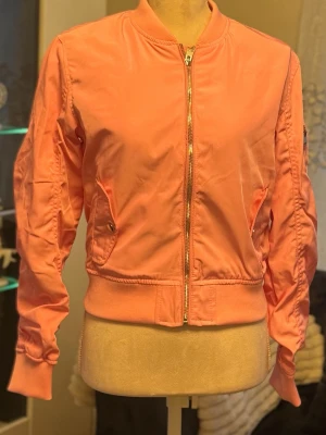 Rosa bomberjacka med dragkedja - Snygg rosa bomberjacka med silvrig dragkedja framtill och ribbade muddar vid ärmslut och midja. Jackan har två fickor med knappstängning och en klassisk ficka på ärmen. Tillverkad i ett glansigt material som ger en fräsch look.