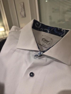 Oscar of Sweden slim-fit vit skjorta, storlek 42 16½ - Klassisk vit skjorta från Orsay i slim fit-modell. Skjortan har mörkblå knappar och ett snyggt paisleymönstrat blått tyg på insidan av kragen. Perfekt för dig som gillar stilrena plagg med en twist.