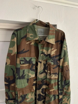 Autentisk vintage militär overshirt / Field jacka - Tjena! Nu finns denna riktigt feta Autentiska vintage militär overshirten / Field jackan till salu! Den är i toppskick utan några direkta skador, speciellt med tanke på åldern av denna jackan. Denna var tillverkad till US marine corps vilket syns med stampningen på bröstet. Och just denna tillverkades troligen runt 1983/84. Jackan har även det klassiska ”woodland” camo mönstret och är i storleken är medium regular. 