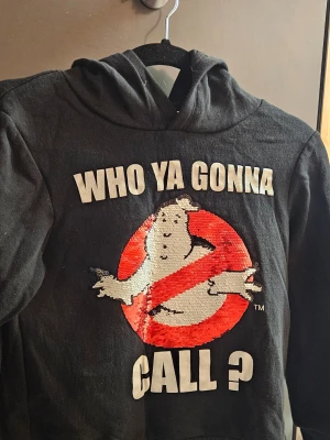Ghostbusters hoodie med paljetter - Svart hoodie med huva och Ghostbusters-tryck framtill. Texten 'WHO YA GONNA CALL?' och den klassiska loggan i paljetter som kan ändra färg när man drar med handen. Perfekt för dig som gillar retro och filmreferenser.