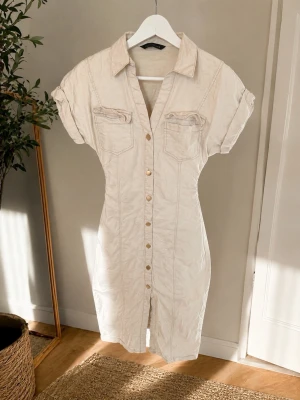 Beige jeansklänning med knappar - Säljer en klänning med klassisk skjortkrage, korta ärmar och dekorativa bröstfickor. Klänningen har knappar hela vägen framtill och markerad midja för en snygg siluett. Perfekt för dig som gillar stilrena och trendiga plagg.