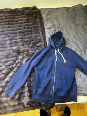 Blå hoodie från Polo Ralph Lauren - Mörkblå hoodie från Polo Ralph Lauren med dragkedja framtill och vit dragsko i huvan. Klassisk design med kängurufickor och liten broderad logga på bröstet. Tillverkad i mjukt bomullsmaterial som känns skönt mot huden.