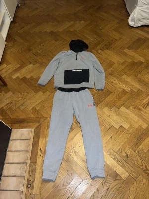 Grått loungewear-set från Under Armour - Snyggt och bekvämt loungewear-set från Under Armour i ljusgrått med svarta detaljer. Setet består av en hoodie med halv dragkedja, svart magficka och logga på bröstet samt matchande byxor med resår i midjan och logga på benet. Perfekt för chill dagar hemma.