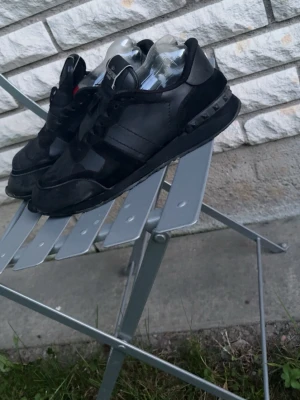 Svarta Valentino sneakers - Snygga helsvarta sneakers från Valentino med klassisk låg siluett. Skorna har detaljer i läder och mocka, samt svarta nitar på hälen för en edgy look. Perfekta för dig som gillar stilrena och exklusiva sneakers med streetkänsla. Båda skorna har hål vid tårna och runtomkring har slitage vilket inte synd där med prisens blir lågt!
