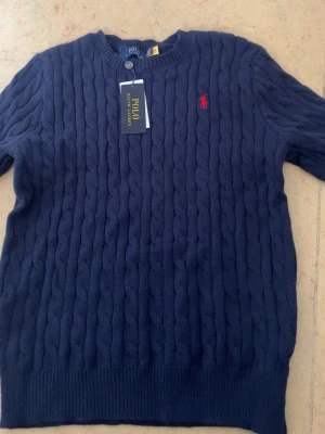 Mörkblå kabelstickad tröja Polo Ralph Lauren - Kabelstickad mörkblå tröja från Polo Ralph Lauren med rund halsringning och röd broderad logga på bröstet. Tröjan har ribbade muddar vid ärmslut och nederkant. Klassisk och stilren design som passar till mycket.