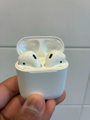 Apple AirPods med laddningsetui - Trådlösa vita Apple AirPods med tillhörande laddningsetui. Hörlurarna har en stilren och kompakt design i blankt plastmaterial och är perfekta för musik, samtal och poddar. Etuiet är smidigt att ta med och laddar hörlurarna automatiskt när de placeras i det.