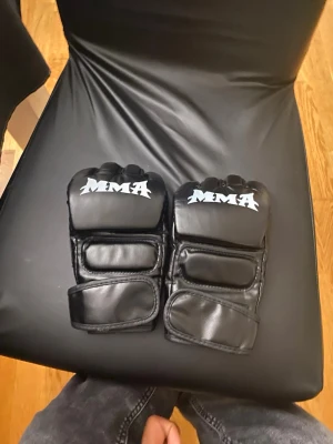 Svarta MMA handskar med öppen hand - Säljer ett par svarta MMA handskar med öppen hand och vadderade knogar. Handskarna har justerbara kardborreband runt handleden och är tillverkade i konstläder. Perfekta för träning eller sparring inom kampsport.