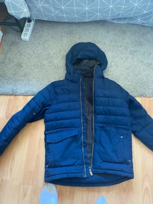 Blå pufferjacka med huva - Snygg blå pufferjacka med huva och dragkedja framtill. Jackan har stora fickor med lock och justerbara ärmslut med kardborre. Perfekt för kyliga dagar och har en modern, avslappnad look.