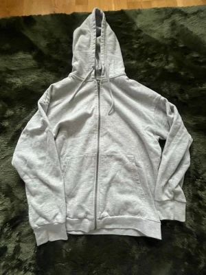 Ljusgrå zip hoodie med huva - En enkel och clean ljusgrå hoodie med dragkedja framtill och stor ficka på magen. Hoodien har justerbar huva med snören och ribbade muddar vid ärmar och nederkant. Perfekt för chill dagar eller när du vill ha en basic look. Har en liten fläck💕