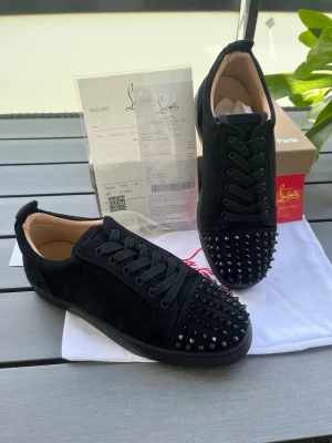 Svarta Louboutin sneakers med spikar - Säljer ett par svarta sneakers från Christian Louboutin i mocka med svarta nitar på tån och klassiska röda sulor. Skorna har svarta skosnören och en låg siluett som ger en clean och edgy look. Perfekta för dig som vill sticka ut med exklusiv streetstyle. Fraktar direkt!