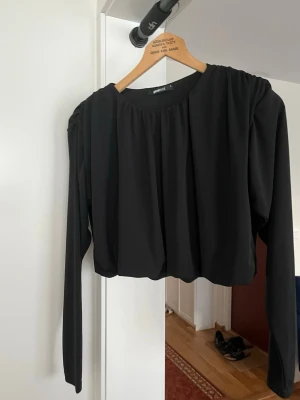 Svart croppad topp från Gina Tricot - Trendig svart croppad topp från Gina Tricot med rund halsringning och långa ärmar. Toppen har en lös passform och lätt rynkade axlar för en cool siluett. Perfekt att styla med höga jeans eller kjol.