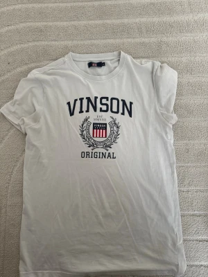 Vit t-shirt från Vinson - Vit t-shirt från Vinson med tryck framtill i marinblått och rött. Klassisk rund halsringning och korta ärmar. Tillverkad i mjuk bomull för en skön känsla. Perfekt för en avslappnad och stilren look.