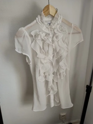 Två vita toppar med volangdetaljer - Säljer två vita toppar från H&M. Ena är en blus med kort ärm, hög krage och volanger framtill. Den andra är en linnetopp med tunna axelband och volang längs urringningen. Båda är perfekta för en chic och feminin look.