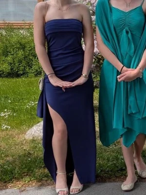 Marinblå axelbandslös balklänning - Säljer en marinblå axelbandslös lång balklänning med slits på sidan. Klänningen har en tight passform och är gjord i ett slätt material som ger en elegant look. Perfekt för dig som vill sticka ut med en stilren och modern klänning. Klänningen har använts en gång så den är i väldigt bra skick.