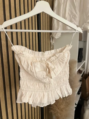Ljusbeige volang topp med knyt - Supersöt ljusbeige linnetopp med bandeau, volangkant nertill och smockad midja. Toppen har en liten knytning framtill och är perfekt för varma dagar. Luftigt material och feminin look.