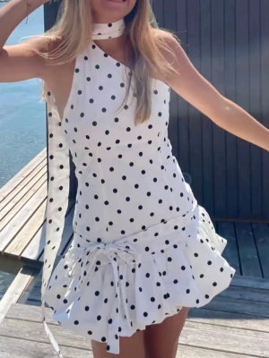 Vit prickig asymmetrisk klänning - Snygg vit klänning med svarta prickar, asymmetrisk axel och volangkant nedtill. Klänningen har en dekorativ knytning i midjan och en matchande scarf runt halsen. Perfekt för sommarens alla tillfällen!