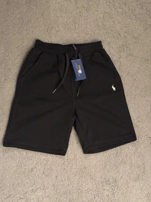 Svarta shorts från Polo Ralph Lauren - Svarta shorts från Polo Ralph Lauren med elastisk midja och snörning. Klassisk logga broderad i vitt på vänster ben. Mjukt material och två sidofickor gör dem perfekta för en avslappnad stil.
