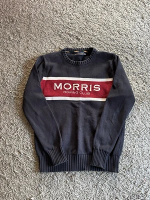 Mörkblå stickad tröja Morris - Stickad tröja från Morris med mörkblå bas, bred vinröd rand och vita detaljer över bröstet. Texten 'Morris Rowing Club' är broderad framtill. Tröjan har rund halsringning och ribbade muddar vid ärmslut och nederkant.
