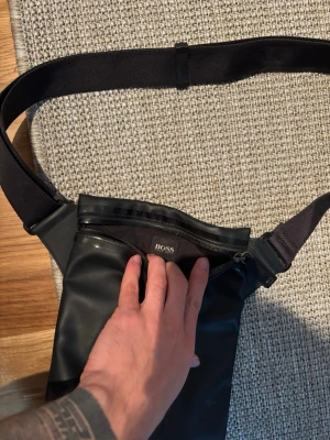 Svart axelväska från BOSS - Snygg svart axelväska från BOSS i slätt material med justerbar axelrem. Väskan har en dragkedja upptill och en diskret BOSS-logga framtill. Perfekt för att bära det viktigaste och ger en clean look till din outfit.