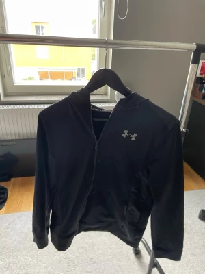 Svart hoodie från Under Armour 147-158 - Snygg svart hoodie från Under Armour med dragkedja framtill och klassisk logga på bröstet. Hoodien har huva och två sidofickor med dragkedja. Tillverkad i ett mjukt och stretchigt material som passar perfekt till både träning och chill.