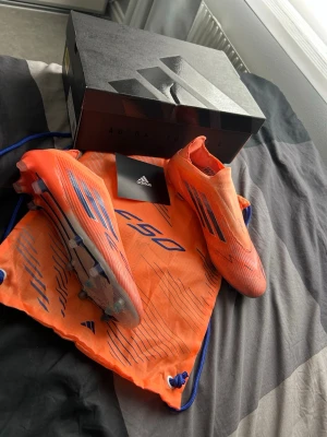 F50 Elite  - Säljer ett par Adidas F50 fotbollsskor i en riktigt snygg orange färg med blå detaljer och klassiska tre ränder på sidan. Skorna har låg vikt, strömlinjeformad design och är tillverkade i syntetmaterial för bästa känsla på planen. Perfekta för dig som vill sticka ut!